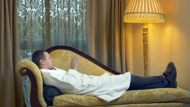 Бизнесмен в халате отдыхает на диване с телефоном вечером на фоне окна — стоковое видео