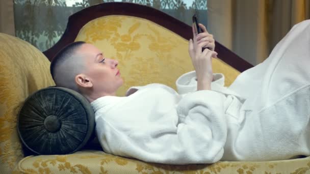 Bela mulher careca em um roupão de banho fica em um sofá com um telefone à noite contra o fundo de uma janela em um interior luxuoso — Vídeo de Stock