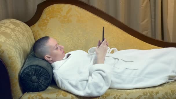 Beautiful bald woman in a bathrobe lies on a sofa with a phone in the evening against the background of a window in a luxurious interior — Stock Video