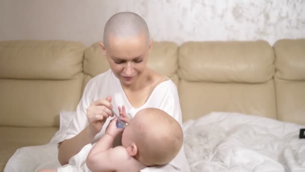 Uma mulher careca segura nos braços e se alimenta com uma colher um bebê de sete meses sentado em um sofá na sala de estar . — Vídeo de Stock