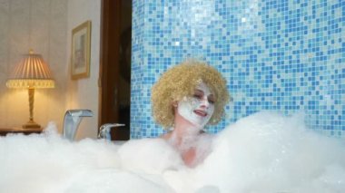 Kafkasyalı neşeli sarışın kadın banyoda spa yapıyor, köpük banyosunda yatıyor, yüzünde beyaz bir maskeyle köpük bulutlarıyla oynuyor.