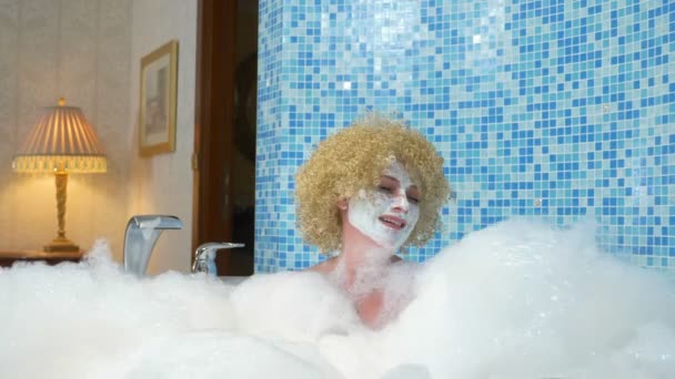 Mulher loira caucasiana alegre goza de um spa em casa no banheiro, encontra-se em um banho de espuma com uma máscara branca em seu rosto, brinca com nuvens de espuma — Vídeo de Stock