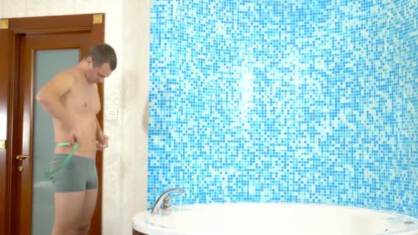 Hombre joven caucásico que mide el cuerpo con cinta métrica mientras está de pie en el baño. concepto humorístico — Vídeo de stock