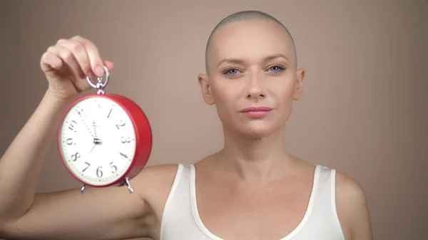 Beautiful bald woman holds an alarm clock and a pink ribbon in her hands. gentle pastel background. — Stock Photo, Image