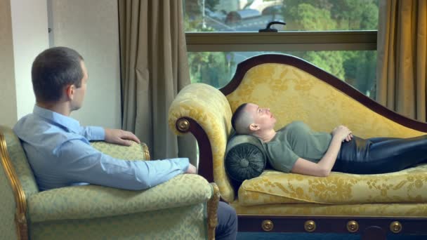 Психолог работает с молодой лысой женщиной, лежащей на диване в стильном кабинете — стоковое видео
