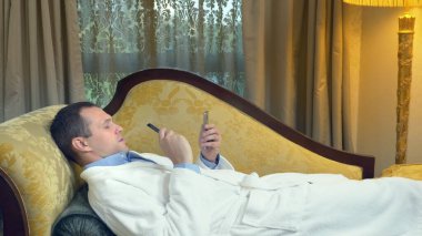 Bornozlu bir iş adamı kanepede telefonla rahatlar ve akşamları bir pencerenin arkasında sigara içer.