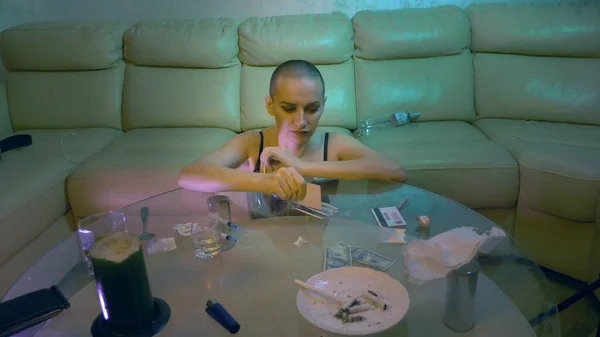 Éjszakai élet koncepció. elbűvölő kopasz nő használ fehér por ül egy üveg asztal alkohol és fecskendők a nappaliban — Stock Fotó