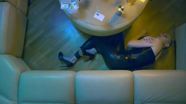 Una mujer calva de lujo en un corsé de cuero negro se encuentra en convulsiones en el suelo contra una mesa con alcohol y jeringas. vista desde arriba. Concepto de sobredosis de drogas, retirada — Foto de Stock