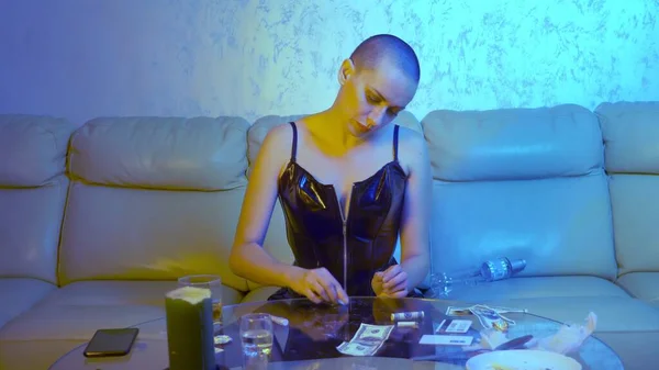 The concept of night life. Glamorous bald woman rubs white powder in gums sitting at a glass table with alcohol and syringes in the living room