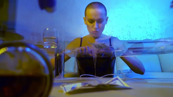 The concept of night life. Glamorous bald woman rubs white powder in gums sitting at a glass table with alcohol and syringes in the living room