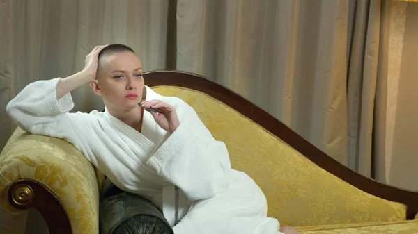 A beautiful bald girl in a bathrobe lies on a luxurious sofa against the background of a window and smokes an electronic cigarette. copy space — Stock Photo, Image