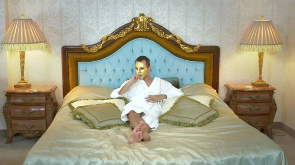 Un hombre guapo con una máscara dorada y un albornoz descansa sobre una lujosa cama fumando un cigarrillo electrónico. mirando a la cámara — Foto de Stock