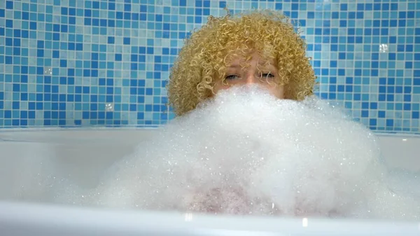 Vidám kaukázusi szőke nő élvezi az otthoni spa a fürdőszobában, fekszik a hab fürdő fehér maszk az arcán, játszik hab felhők — Stock Fotó