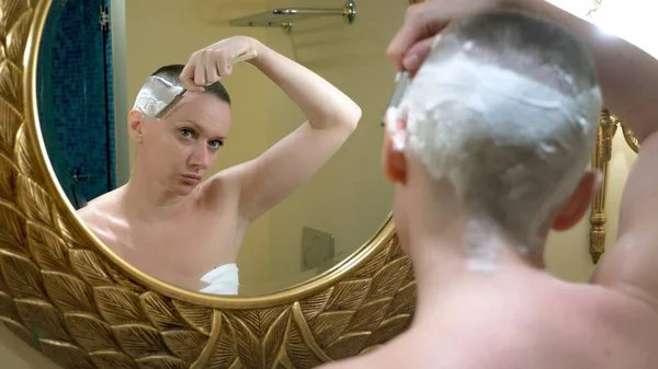 Mooie kale vrouw kijkt in een luxe badkamer spiegel en doet haar haar, scheert haar haar met een gevaarlijk scheermes — Stockfoto