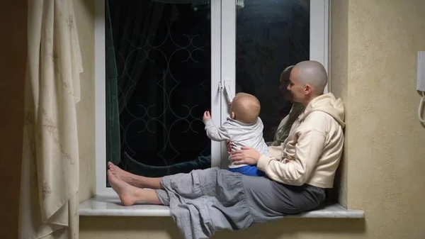 Φαλακρή γυναίκα και το μωρό κάθονται μαζί στο παράθυρο το βράδυ. — Φωτογραφία Αρχείου
