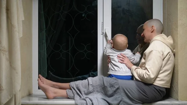 Kale vrouw en baby zitten samen aan het raam in de avond. — Stockfoto