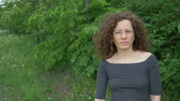 Une femme douteuse avec les cheveux bouclés dans les lunettes se tient près d'un arbre — Video