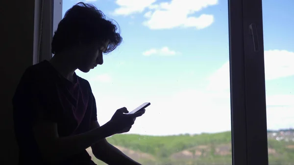 Silhueta. O menino usa um telefone, pela janela contra o céu azul e colinas — Fotografia de Stock