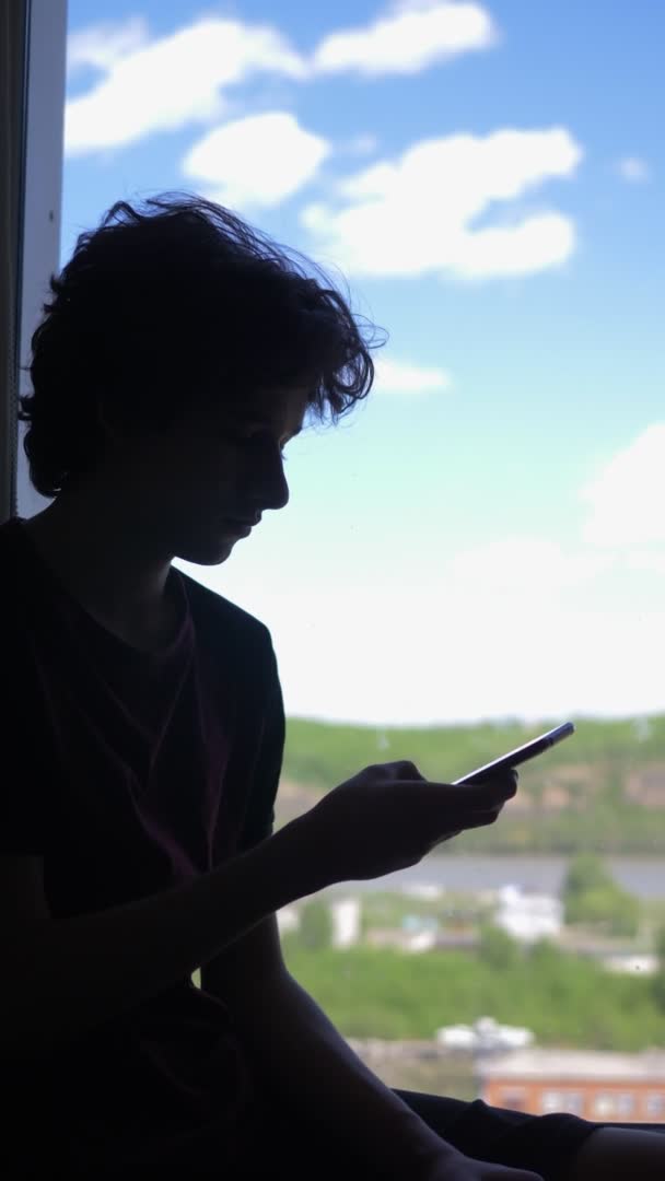 Vertikal. Silhouette. Der Junge benutzt ein Telefon, am Fenster gegen den blauen Himmel — Stockvideo