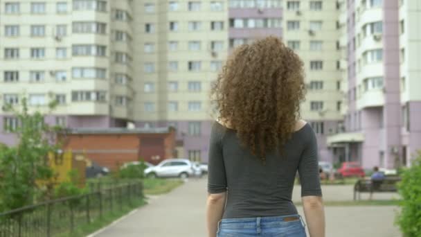 Vista para trás. mulher com cabelo encaracolado caminha ao longo de uma rua da cidade, o vento em seu cabelo — Vídeo de Stock