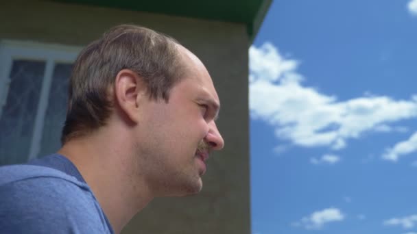 Homem careca senta-se ao ar livre contra o fundo de uma casa de campo e céu — Vídeo de Stock