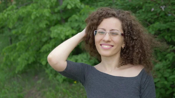 Mooi meisje in bril met krullend haar wandelingen in het park op een achtergrond van bomen — Stockfoto