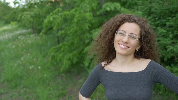 Красивая девушка в очках с вьющимися волосами гуляет по парку на фоне деревьев — стоковое фото