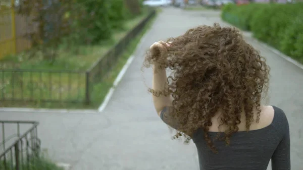 Vista trasera. mujer con el pelo rizado toca su cabello de pie en una calle de la ciudad —  Fotos de Stock