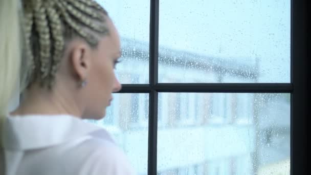 Portret. fata cu împletituri afro stă acasă lângă fereastră, ploaie pe stradă — Videoclip de stoc