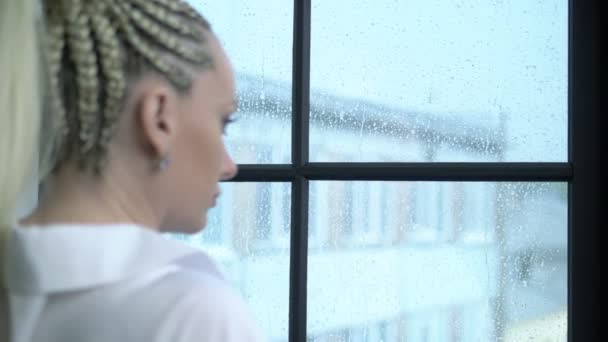 Retrato. chica con trenzas afro se sienta en casa junto a la ventana, lluvia en la calle — Vídeos de Stock