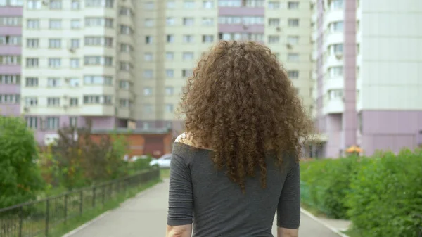 뒷 쪽을 봐. 머리가 곱슬곱슬 한 여자 가 도시의 거리를 걷고, 그녀의 머리카락에 바람 — 스톡 사진