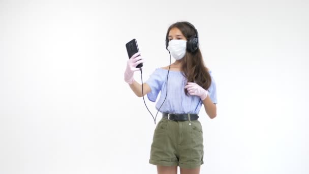 Odizolowane białe tło. piękna dziewczyna w rękawiczkach i maska medyczna w słuchawkach sprawia selfie na smartfonie — Wideo stockowe