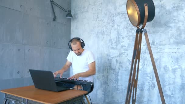 Un hombre con auriculares escribe música electrónica con un teclado midi y un portátil — Vídeo de stock