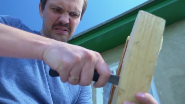 Kale man met snor snijdt een gat in een houten plank met een mes buiten — Stockvideo