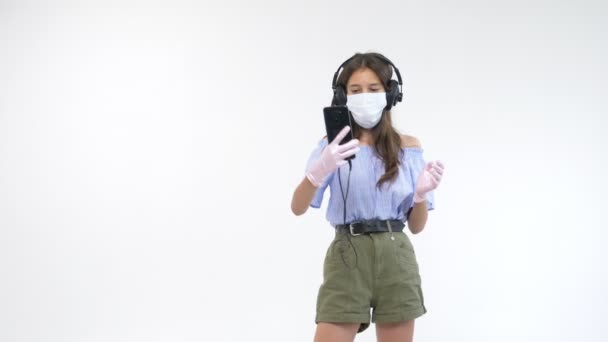 Bílé pozadí. dívka v rukavicích a lékařská maska ve sluchátkách dělá selfie — Stock video