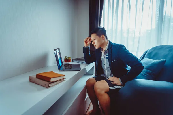 Young Asian Businessman wear Formal Suit with Short Pants Having Video Conference Meeting with Business Partner on Sofa at Home in Living Room during Quarantine Period.