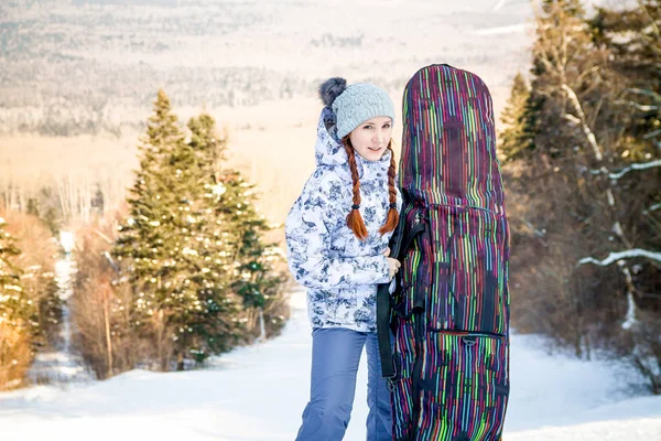 Girl snowboarder standing on a ski track, holding a case with her snowboard and enjoying the ski resort