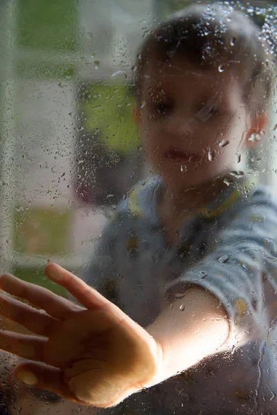 sad portrait of a boy through the glass with drops. Child touches glass.