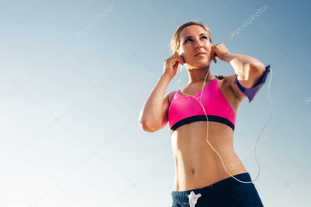 low angle view of sportswoman with smartphone in running armband case putting earphones against blue sky 