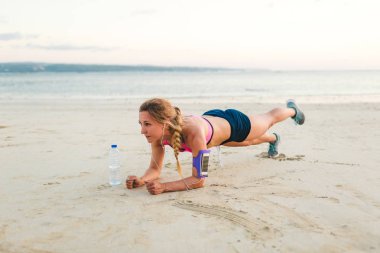 omuz çantası ve şişe suyu kum plajındaki tahta yapmaya çalışan smartphone ile kulaklıklar içinde genç sporcumuz 