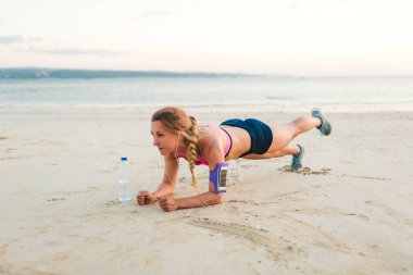 young sportswoman in earphones with smartphone in running armband case and bottle of water doing plank on sandy beach  clipart