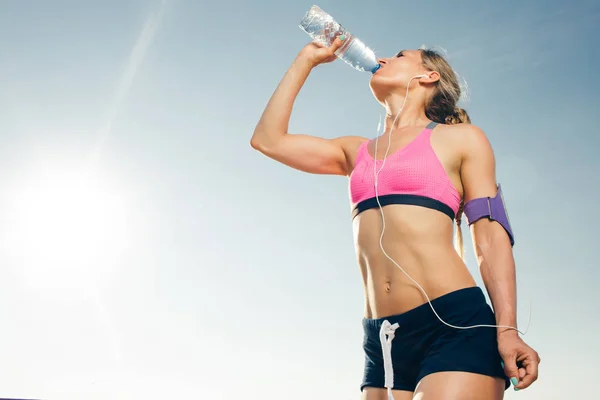 智能手机耳机中年轻运动员的低角观在运行臂章案例饮用水从瓶子对蓝天 — 图库照片
