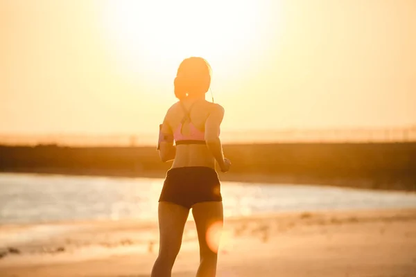 日光に対してビーチでジョギング ランニングのアームバンド ケースにスマート フォンをイヤホンでスポーツウーマンのシルエットの背面図 — ストック写真