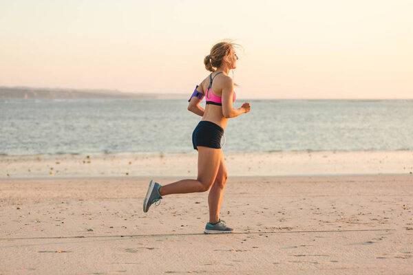 side view of sportswoman in earphones with smartphone in running armband case jogging on beach with sea behind