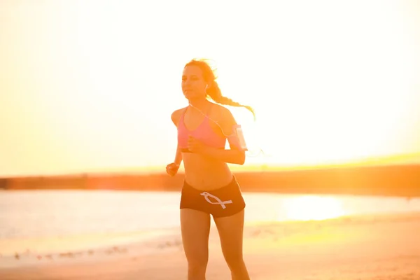 silhouette of sportswoman in earphones with smartphone in running armband case jogging on beach against sunlight