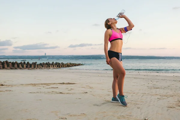 年轻的运动员在耳机与智能手机在臂章案例饮用水从瓶在海滩与海后面 — 图库照片
