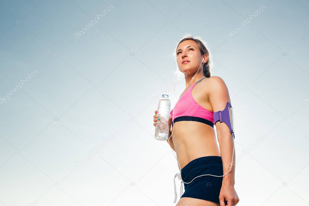 young sportswoman in earphones with smartphone in running armband case holding bottle of water against blue sky