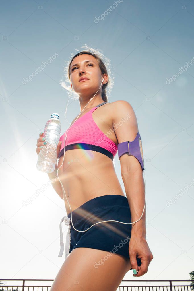 low angle view of young sportswoman in earphones with smartphone in running armband case holding bottle of water against blue sky