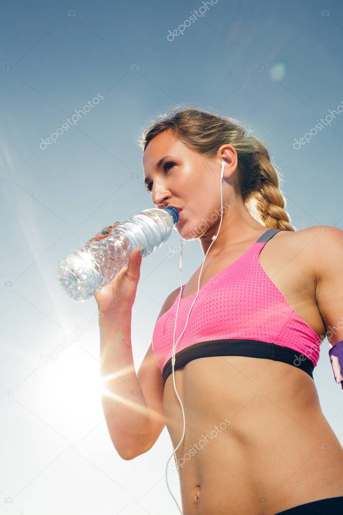 low angle view of young sportswoman in earphones drinking water from bottle against blue sky