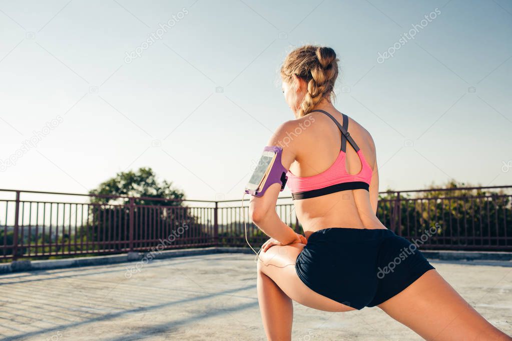 rear view of sportswoman in earphones with smartphone in running armband case stretching on rooftop 